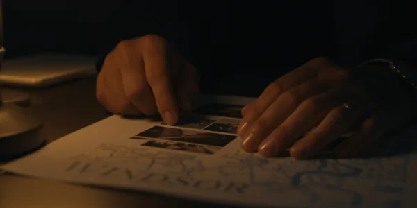 Close-up of investigator's hands reviewing surveillance photos on dark desk, single warm desk lamp creating dramatic shadows, Windsor city map partially visible