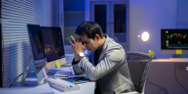Stressed investor sitting at a desk in front of multiple computer monitors displaying stock market charts, appearing concerned about financial losses or investment fraud