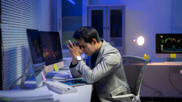 Stressed investor sitting at a desk in front of multiple computer monitors displaying stock market charts, appearing concerned about financial losses or investment fraud